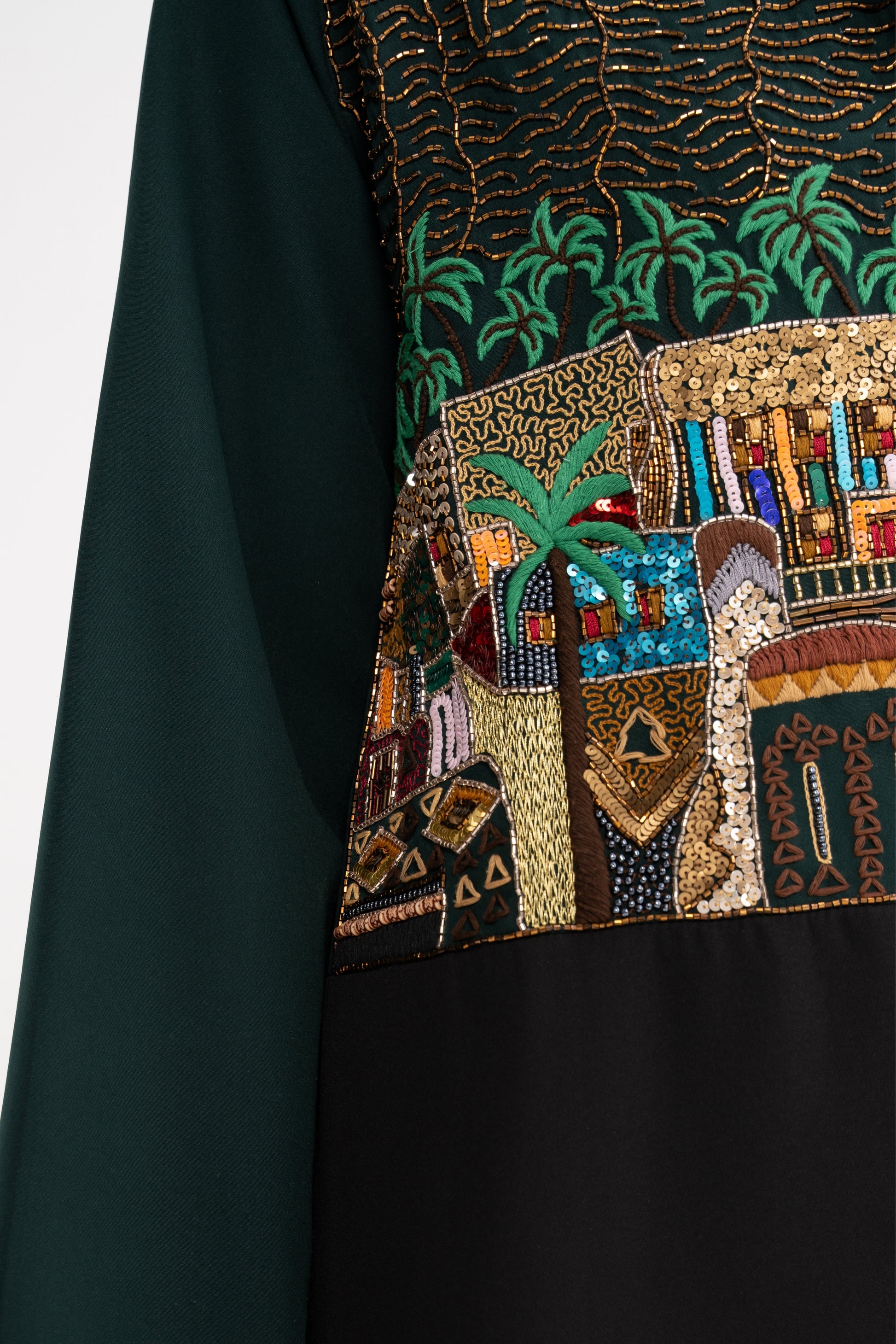 Close-up of a dark green fabric with intricate beadwork and embroidery featuring architectural elements and palm trees.